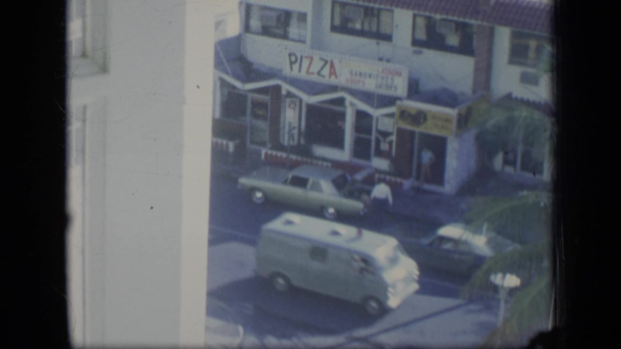 Upper-window view of a pizza storefront, parked cars, a white van, and street traffic.