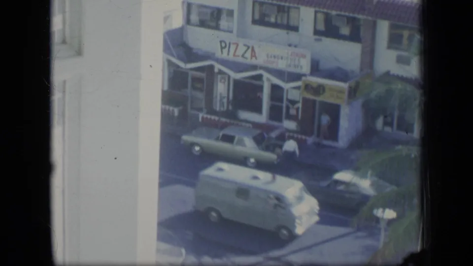 1970 Puerto Rico street view from a window, with a pizza shop sign, older cars, a white van, and pedestrians near the storefront.