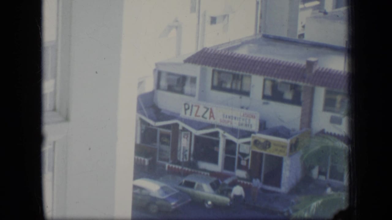 Pizza storefront across a sunny street with cars and a window frame in the foreground.