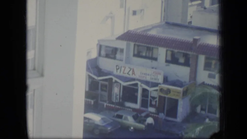 1970 Puerto Rico window-framed view of a pizza shop across the street, with older cars and small figures near the entrance.