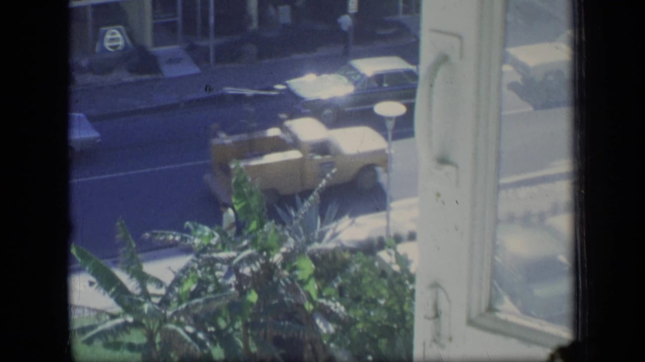 1970 Puerto Rico 8mm frame looking from an upper window toward cars, tropical plants, and a street-level storefront.