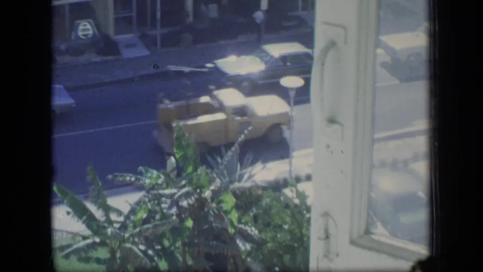 1970 Puerto Rico 8mm frame looking from an upper window toward cars, tropical plants, and a street-level storefront.