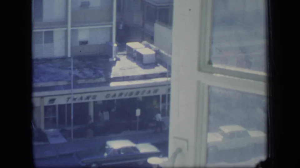 1970 Puerto Rico 8mm frame looking down past a window toward a storefront, sidewalk, cars, and building facade.