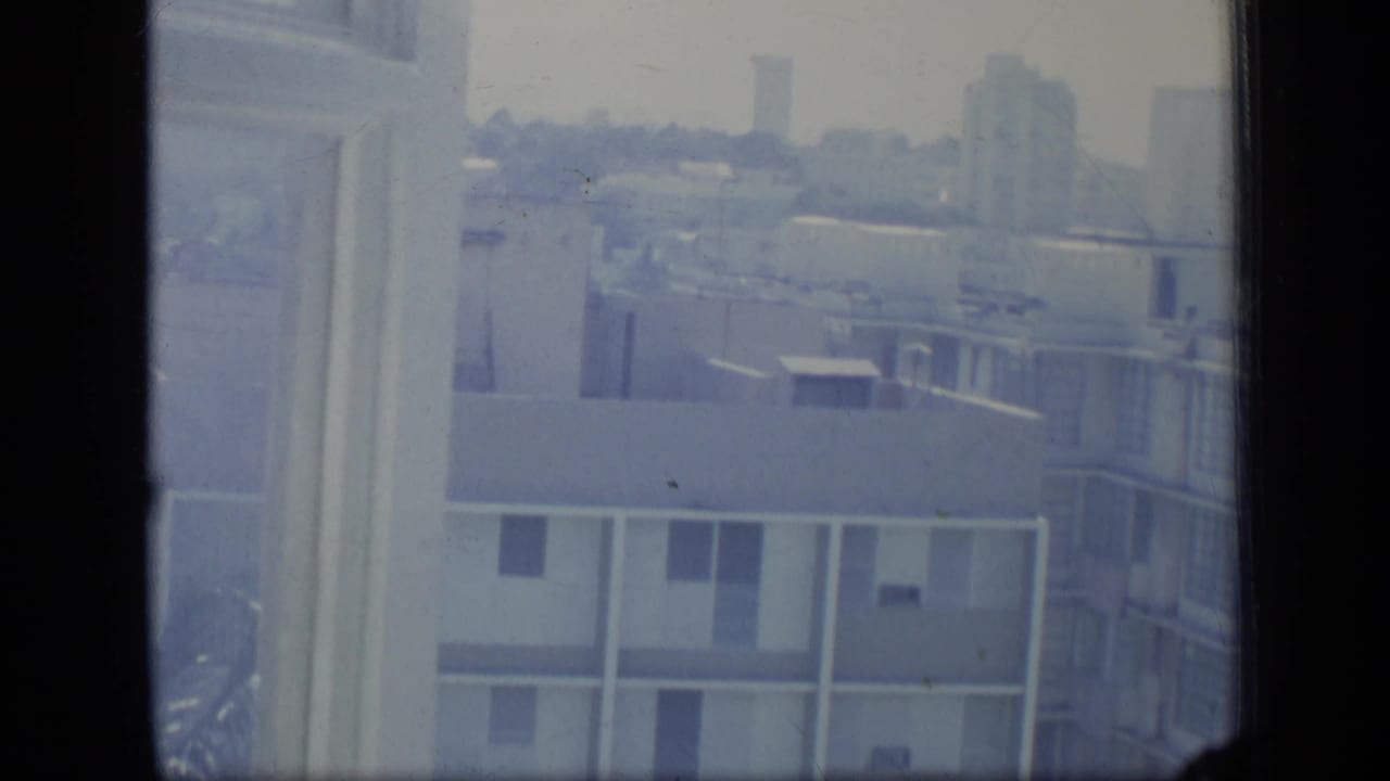 Apartment block and distant buildings viewed beside a window frame.