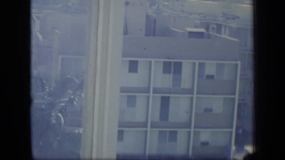 Blue-tinted 8mm frame of Puerto Rico apartment buildings with a window edge and palm fronds near the lower left.