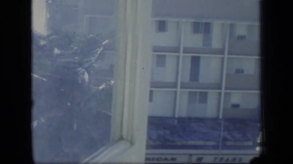 1970 Puerto Rico frame showing a pale apartment facade beyond a window, with haze, film grain, and a partial awning below.