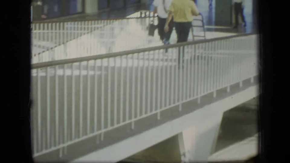 1970 Puerto Rico urban frame showing a white pedestrian bridge, railings, and adults walking near a glass-fronted building.