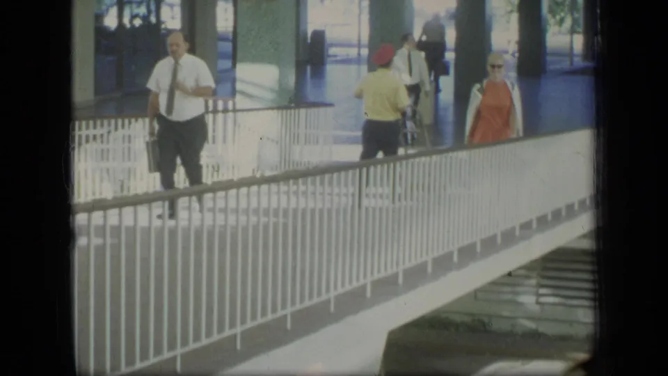 1970 Puerto Rico frame showing adults on a white pedestrian bridge, including business attire, a red hat, and an orange dress.
