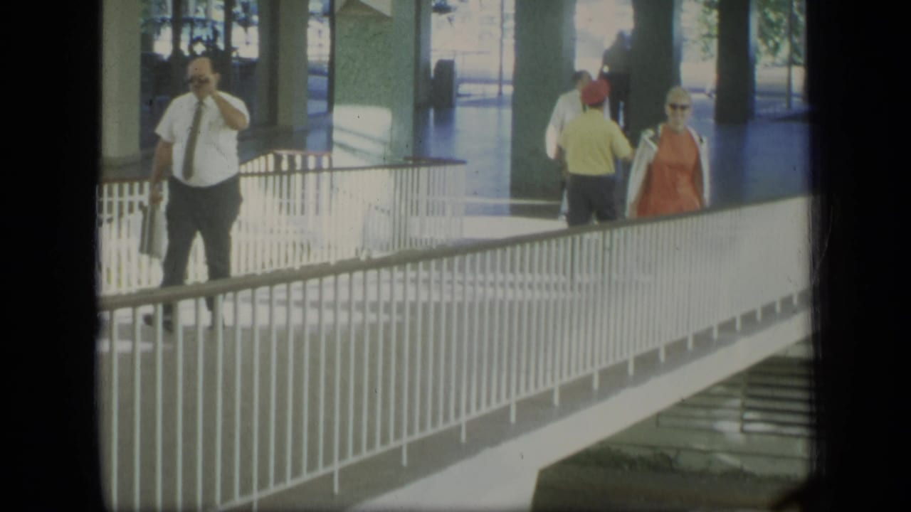 1970 Puerto Rico home-movie frame showing pedestrians in a covered modernist walkway, including a man with a briefcase and a woman in orange.