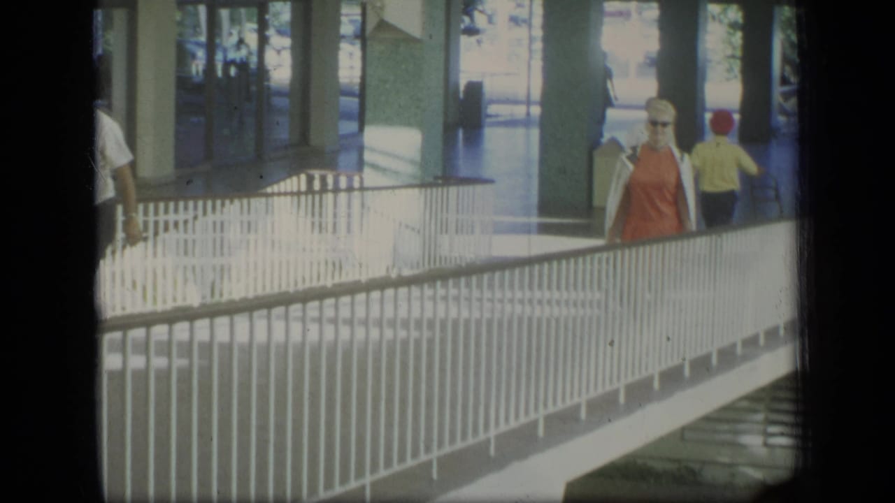 1970 Puerto Rico frame with a woman in an orange dress and sunglasses walking through a covered pedestrian area beside white railings.