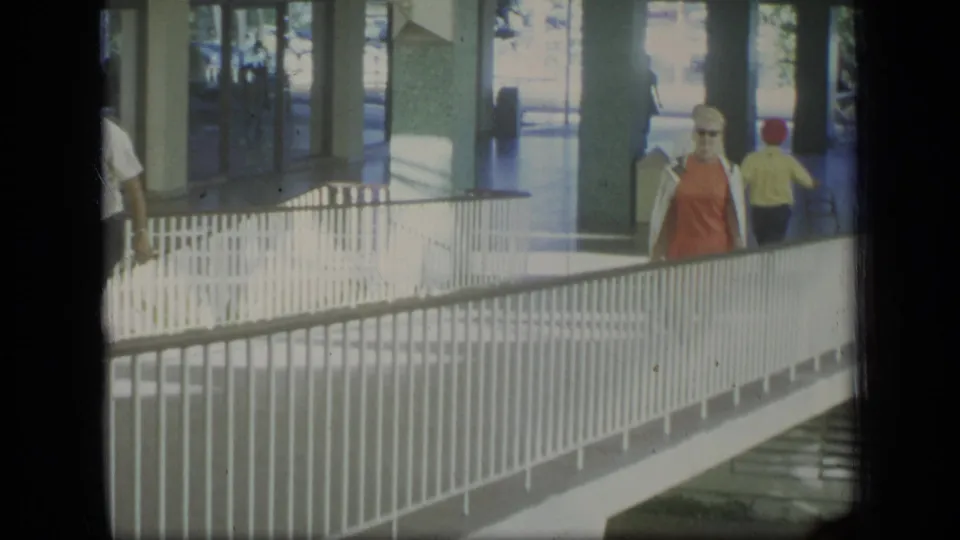 1970 Puerto Rico frame with a woman in an orange dress and sunglasses walking through a covered pedestrian area beside white railings.