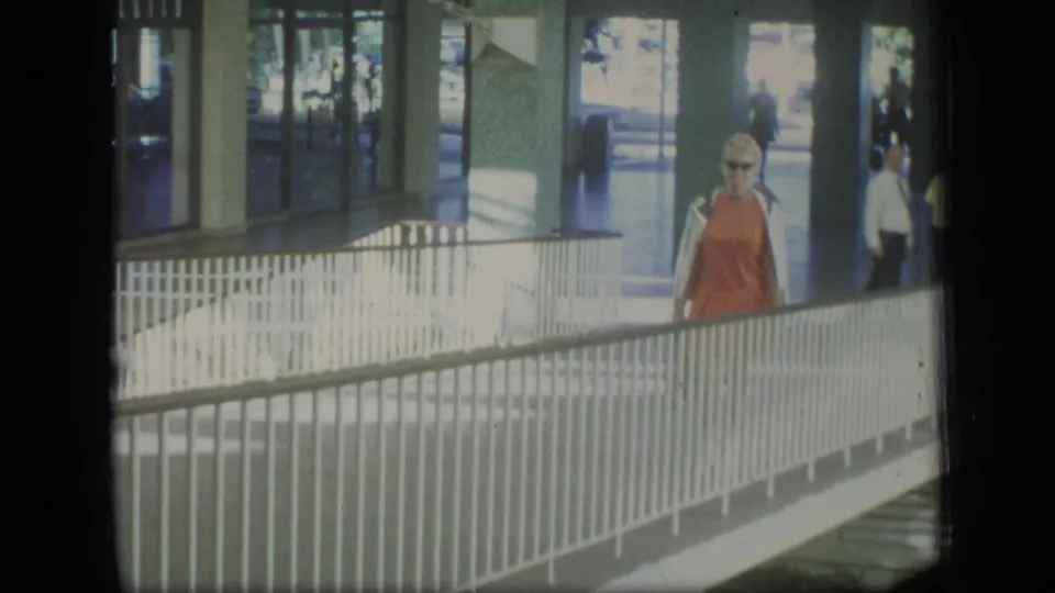 1970 Puerto Rico home-movie frame of a woman in an orange dress approaching along a white-railed covered walkway.