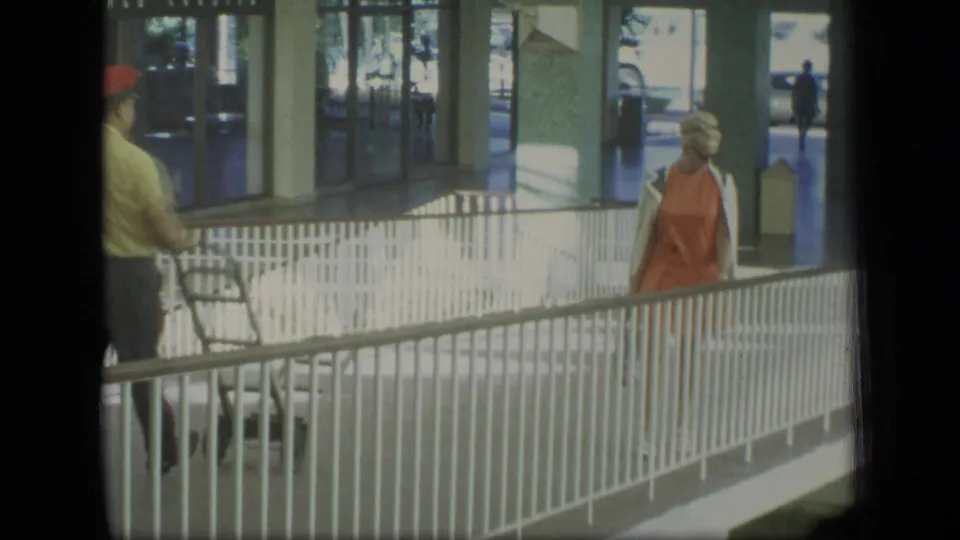 1970 Puerto Rico frame showing a woman in orange near white railings and a worker in a red cap pushing equipment in the foreground.