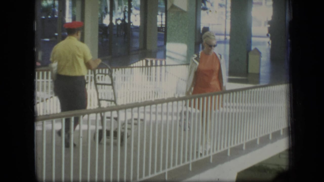 In 1970 Puerto Rico home-movie footage, a woman in an orange dress and sunglasses walks beside a white railing in a covered walkway.