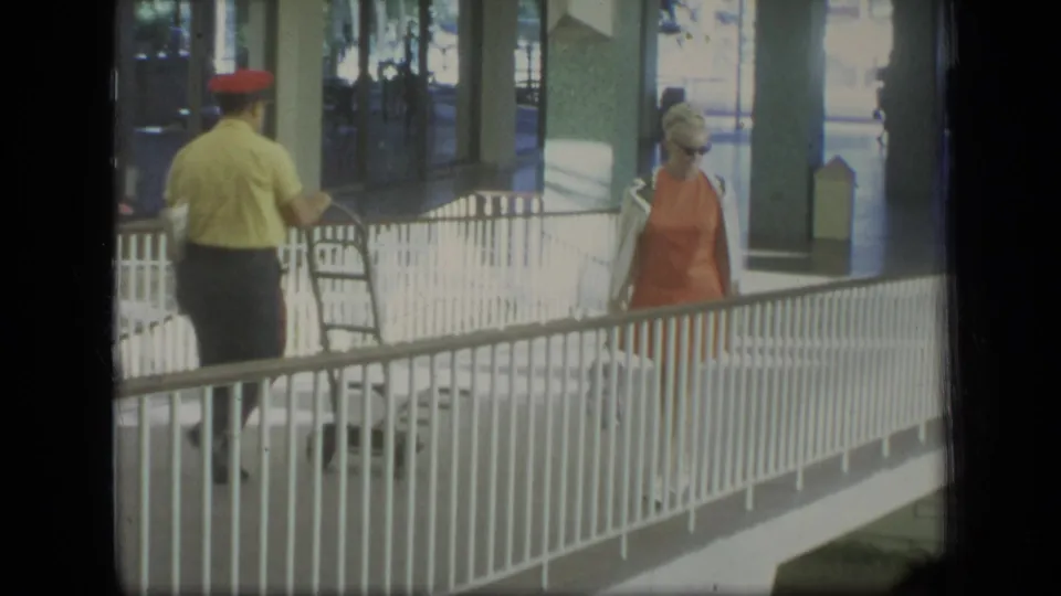 In 1970 Puerto Rico home-movie footage, a woman in an orange dress and sunglasses walks beside a white railing in a covered walkway.