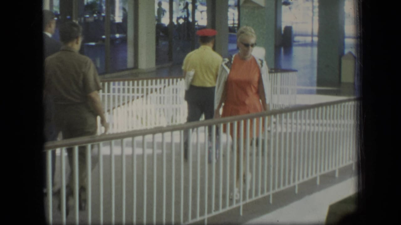Woman in orange dress approaches the camera near a white railing while other pedestrians walk away.