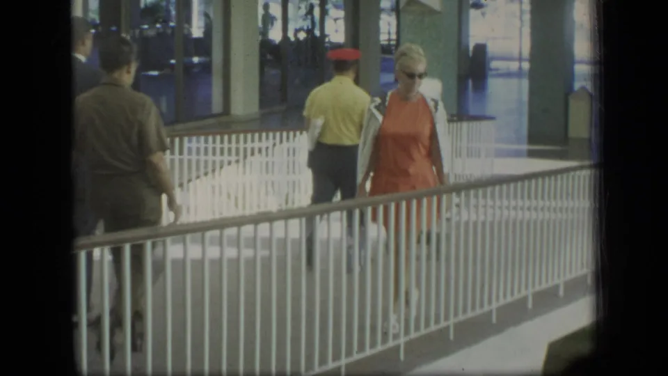 The woman in orange continues along the covered Puerto Rico walkway, with travelers and a luggage cart visible behind her.