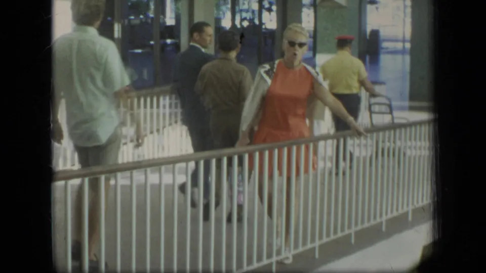 A 1970 Puerto Rico travel scene shows the woman in orange beside the railing, with suited and casually dressed travelers in the background.