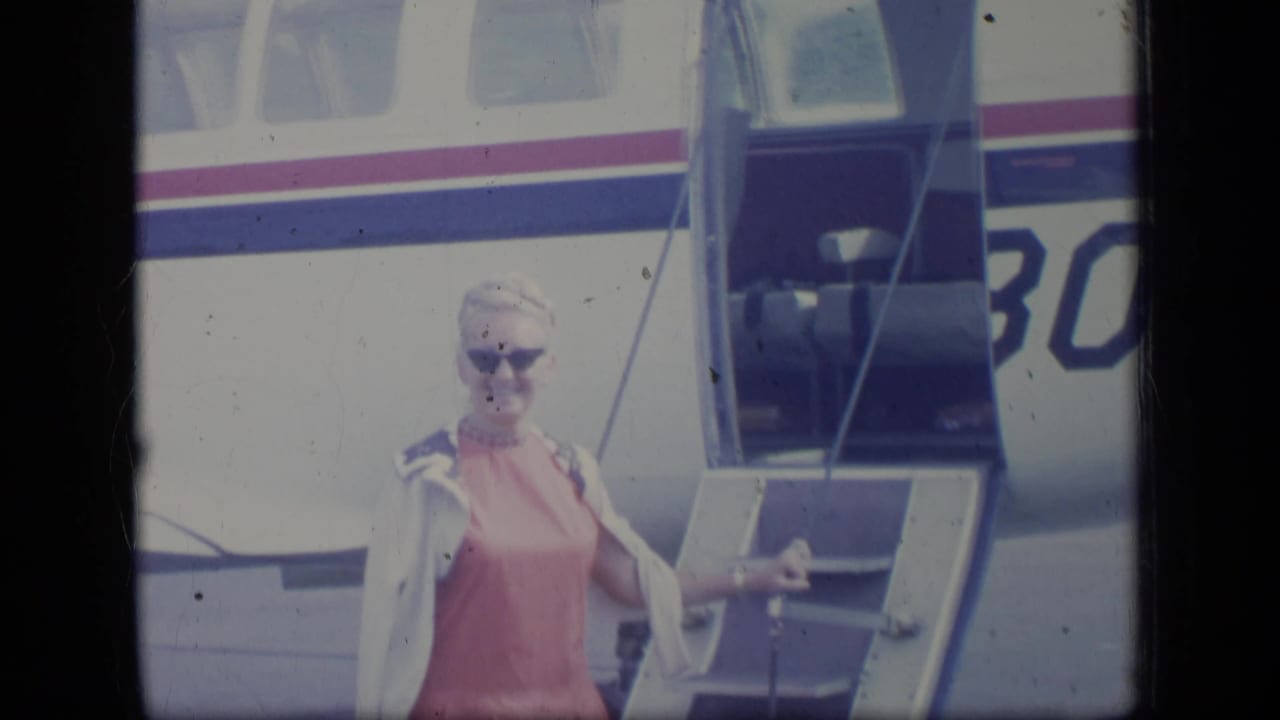 In Puerto Rico 1970 footage, a woman in sunglasses and a pink dress stands on the stairs of a small striped airplane.