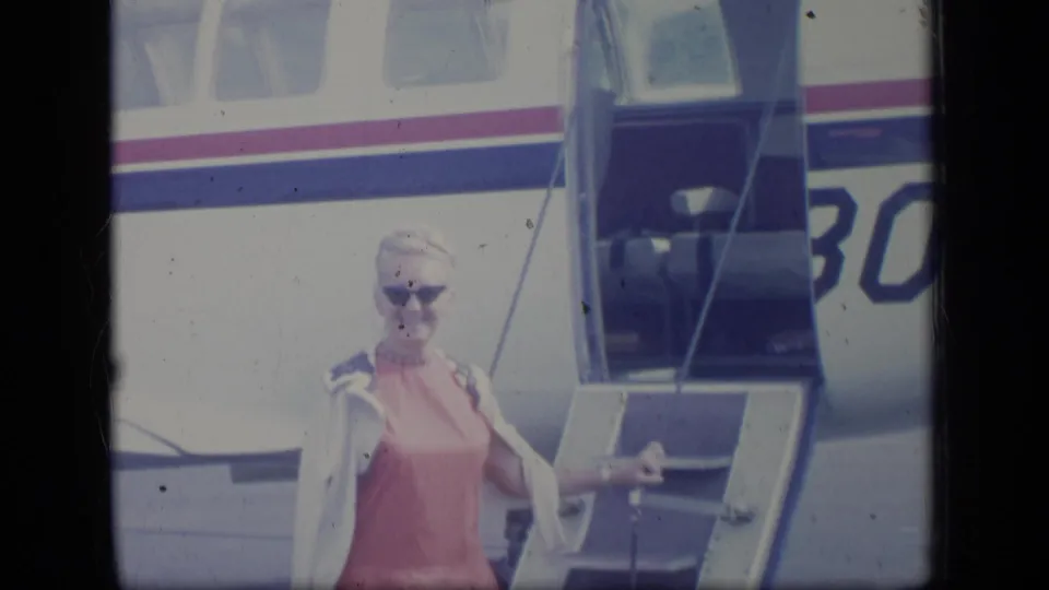 In Puerto Rico 1970 footage, a woman in sunglasses and a pink dress stands on the stairs of a small striped airplane.