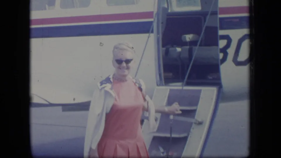 A woman in sunglasses and a pink dress steps down from a small aircraft in 1970 Puerto Rico home-movie footage.