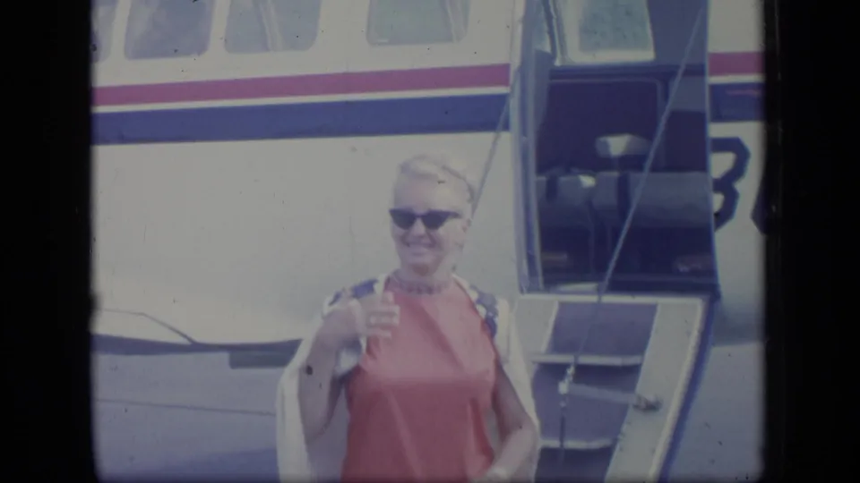 The woman smiles near the open airplane stairs, wearing sunglasses and a pink dress in the Puerto Rico 1970 home movie.