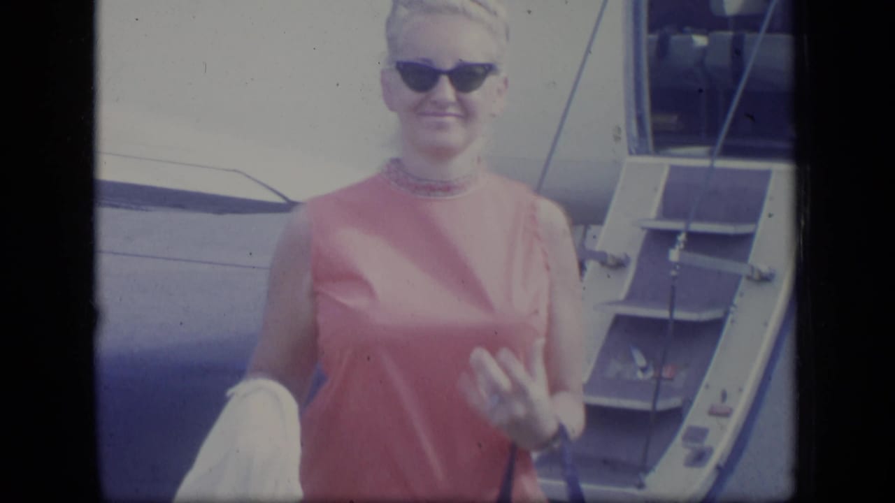 The woman faces the camera beside the aircraft doorway, with sunglasses, a pink dress, and a light garment over one arm.