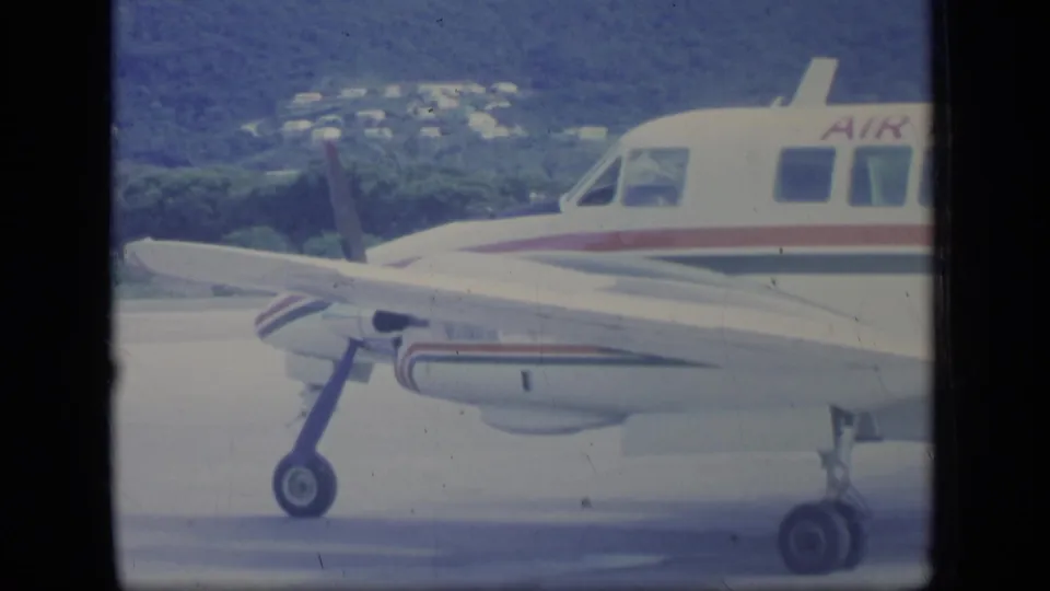 A small Air Indies aircraft with red and blue striping is seen on the tarmac against a green Puerto Rico hillside.
