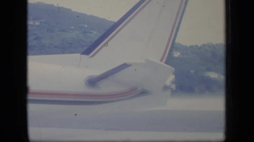 1970 Puerto Rico home-movie frame showing an airplane tail with red and blue stripes above hazy green hills.
