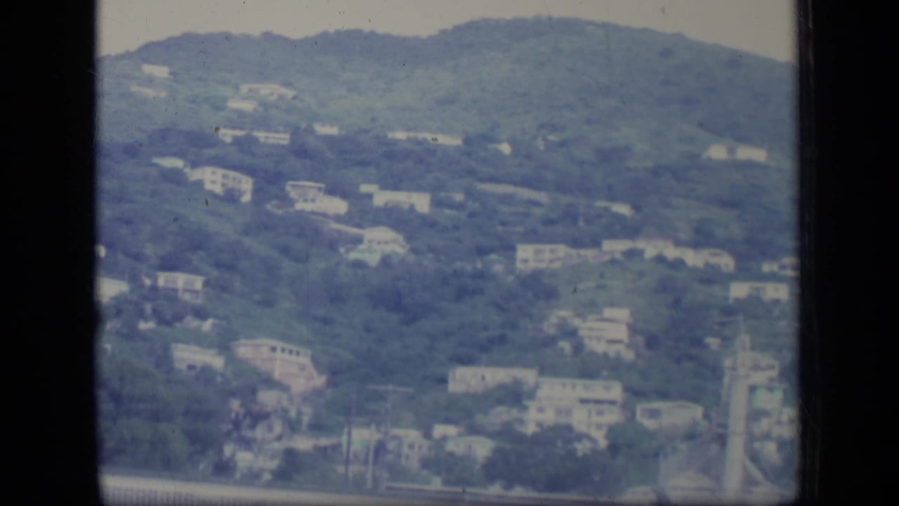 Aerial 8mm view over Puerto Rico with white buildings scattered across a lush hillside under haze.