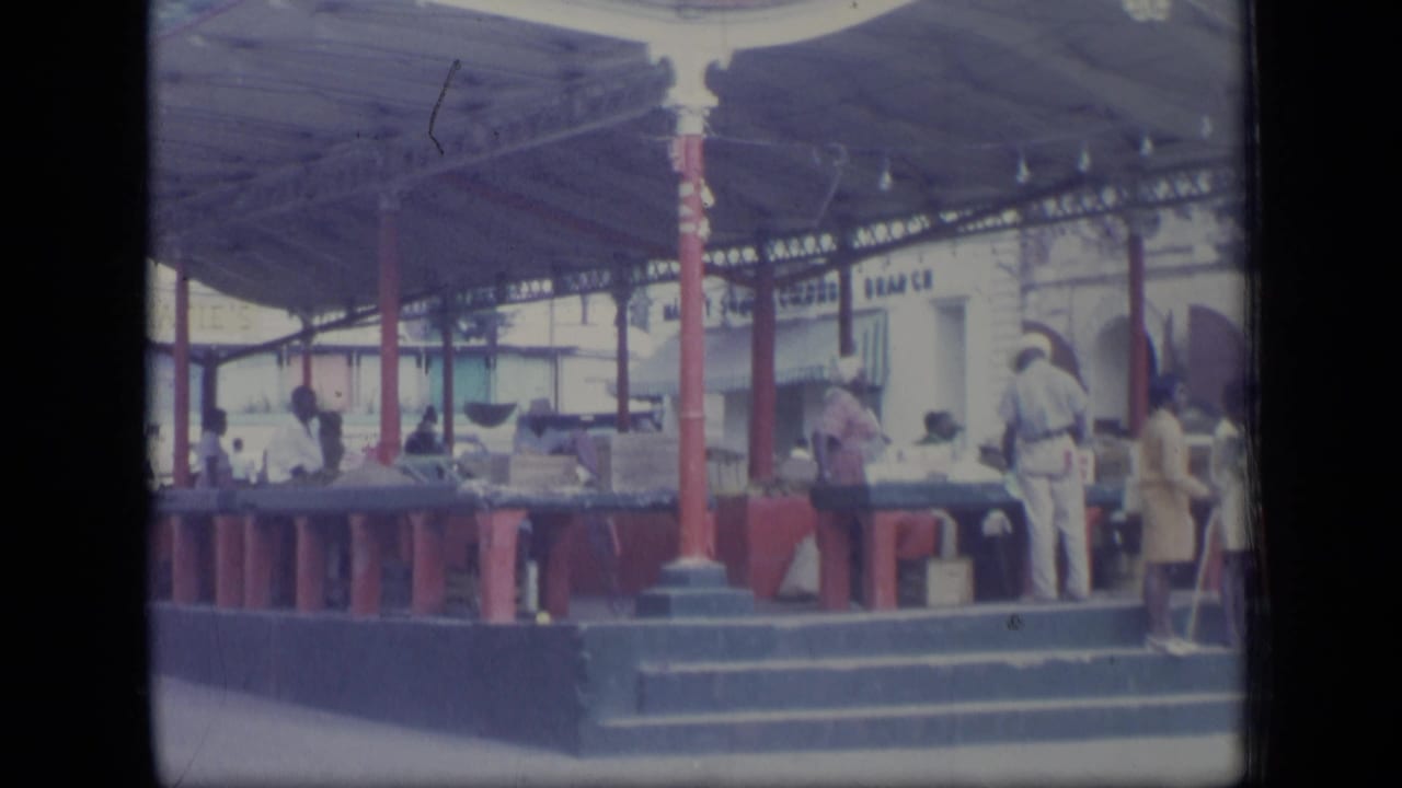 Covered market with vendors and shoppers around counters under red posts.