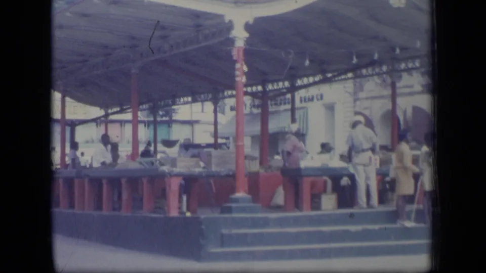 1970 Puerto Rico home-movie frame showing people at a covered market with red posts, counters, steps, and pale buildings behind.
