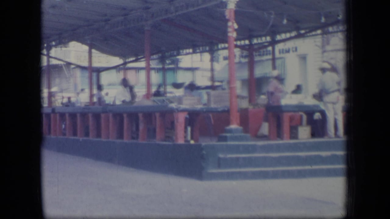 1970 Puerto Rico market scene with people standing behind and beside counters under a roof supported by red posts.