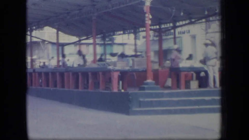 1970 Puerto Rico market scene with people standing behind and beside counters under a roof supported by red posts.