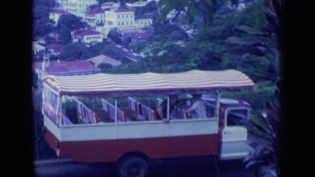 1970 Puerto Rico 8mm frame of an open-sided tour bus with a striped canopy and passengers, backed by hillside buildings and greenery.