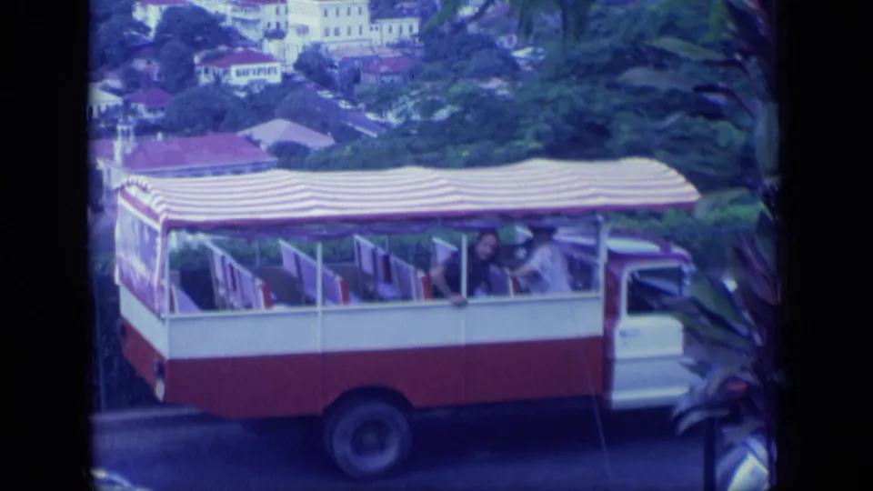 1970 Puerto Rico 8mm frame showing the red and white tour bus near the camera, passengers visible beneath the canopy.