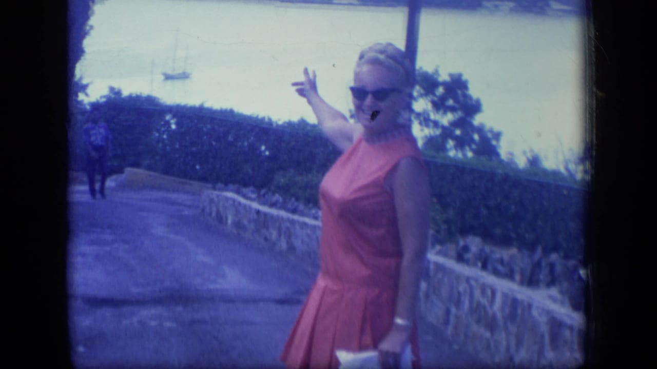 Woman in a red dress and sunglasses points toward a distant blimp above a coastal path.