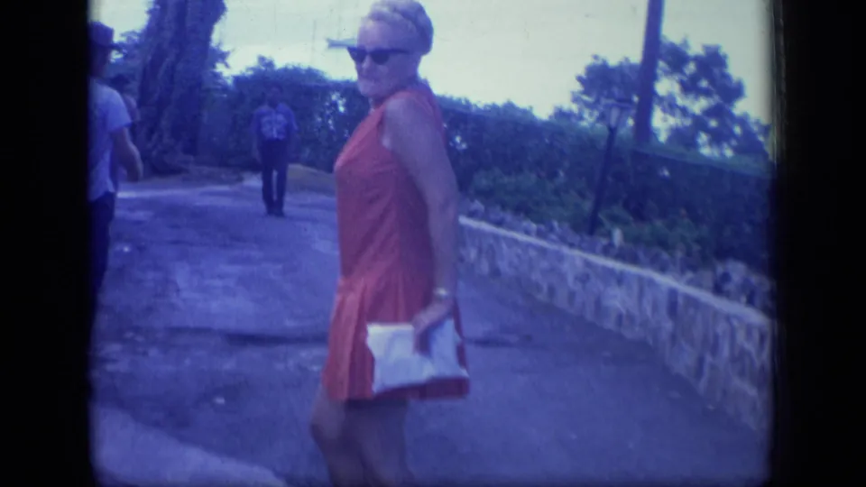A 1970 Puerto Rico travel shot shows the woman in sunglasses walking beside a low stone wall, with another pedestrian in the background.