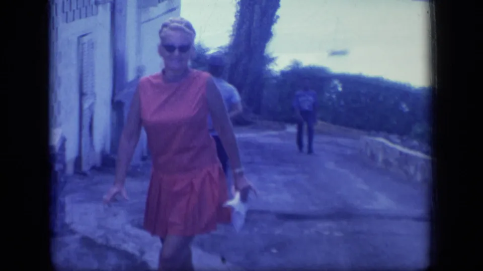 The woman walks toward the camera near a light-colored building, with a stone wall, greenery, and a distant blimp behind her in Puerto Rico.
