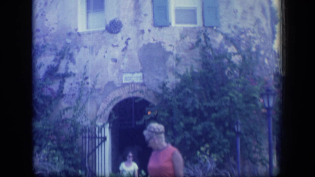 Stone building entrance with greenery and a woman in a red top walking past.