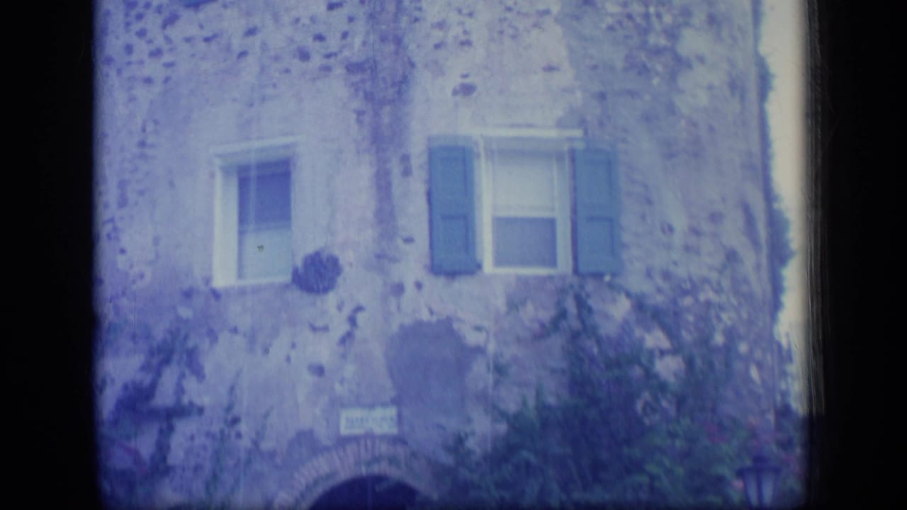 1970 Puerto Rico home-movie frame of a weathered stone building with blue shutters, an arched entry, and plants near the facade.
