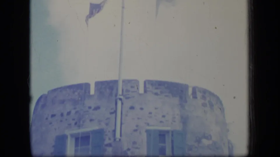 1970 Puerto Rico 8mm frame capturing the top of a weathered stone building with windows and a vertical pole or pipe against a bright sky.