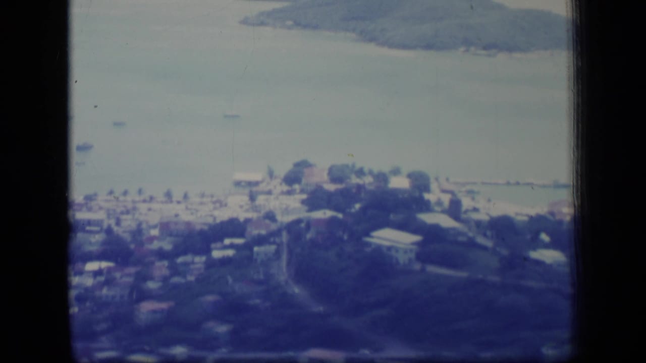 1970 Puerto Rico 8mm frame showing a soft aerial view of shoreline buildings, bay water, and a wooded mountain or island beyond.
