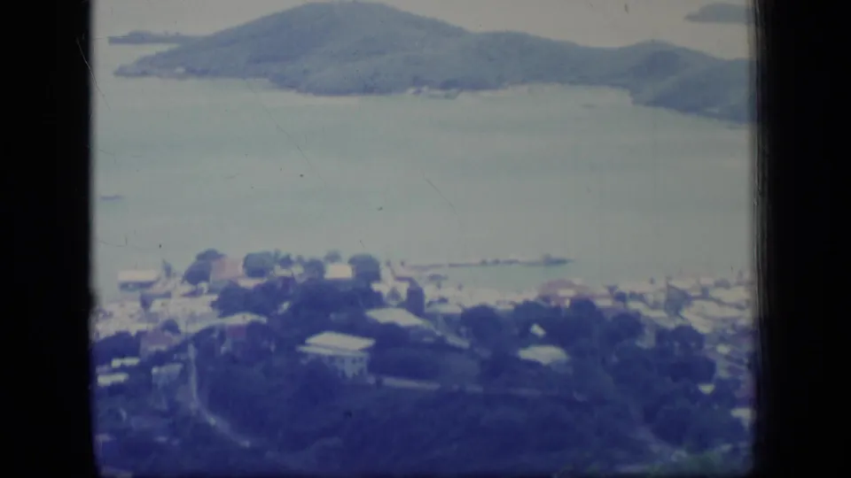 Aerial Puerto Rico home-movie view of clustered shoreline buildings, calm water, small boats, and a wooded mountain in the distance.