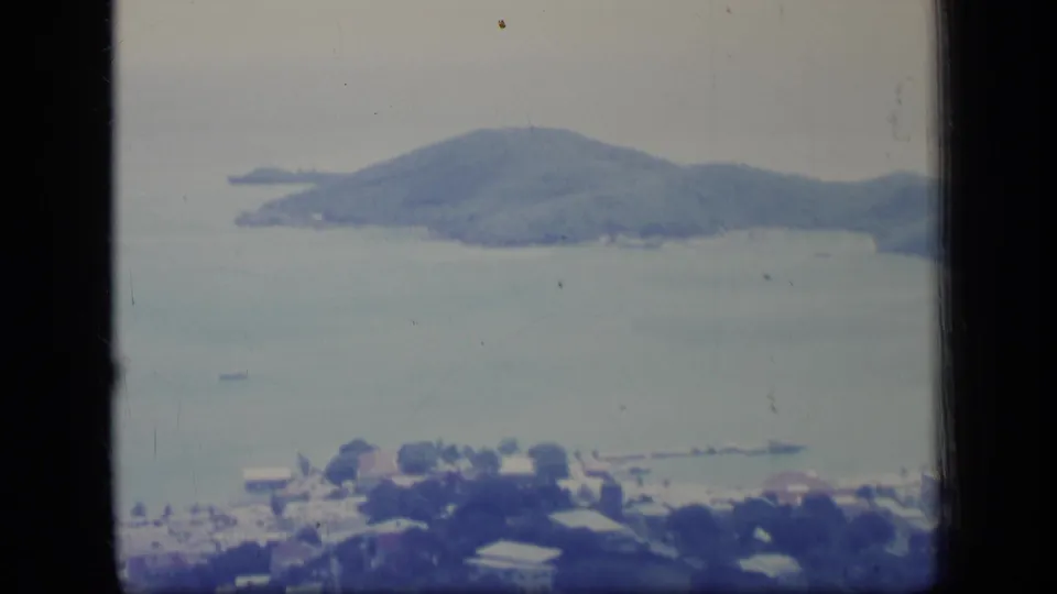 1970 Puerto Rico aerial frame with town rooftops near the lower edge, boats on calm water, and a wooded hill or island beyond.