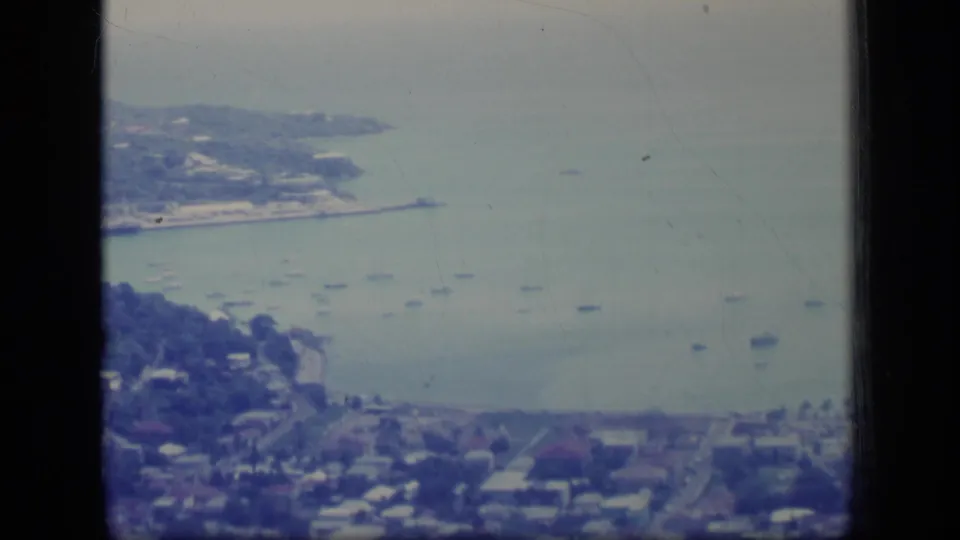 1970 Puerto Rico home-movie frame showing a coastal headland, blue-green bay, small boats, hillside homes, and streets below.