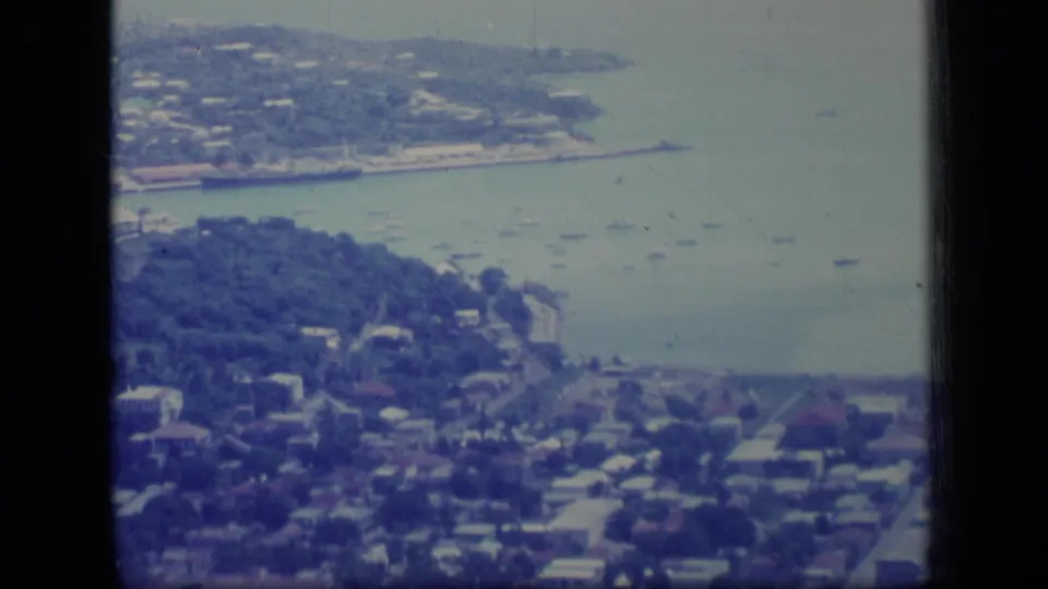 High 1970 Puerto Rico view over wooded residential slopes, rooftops, shoreline roads, and boats anchored in the bay.