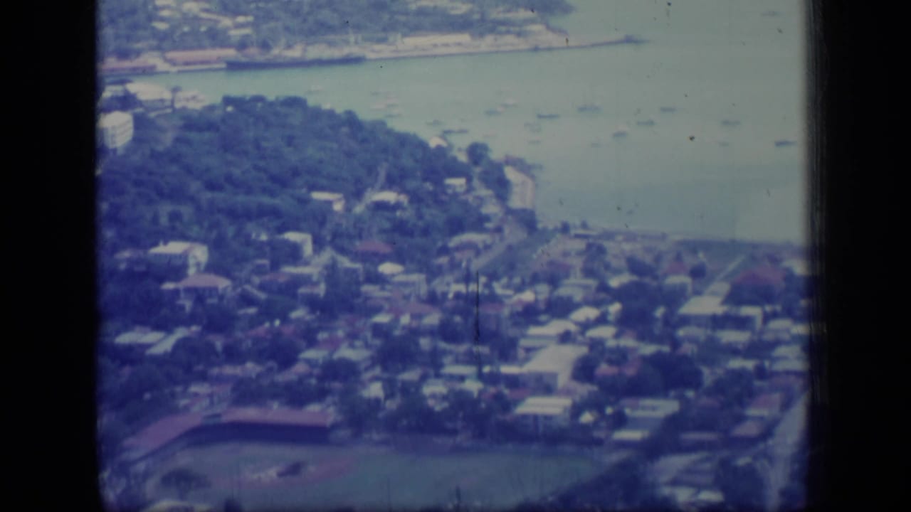 1970 Puerto Rico aerial home-movie frame showing a coastal suburb, bay water, clustered houses, trees, and part of a baseball field.