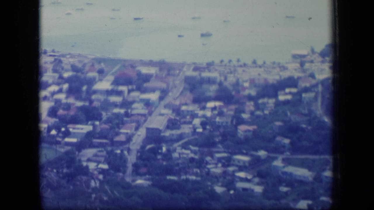 1970 Puerto Rico 8mm aerial frame showing a coastal town, shoreline buildings, boats on calm water, and roads leading inland.