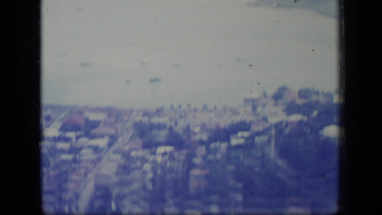 Soft 8mm aerial view in Puerto Rico, with coastal buildings, a road grid, pale water, and small boats in the distance.